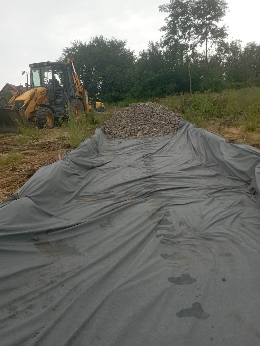 Przygotowanie podłoża w Giebułtowie: rozłożona geowłóknina i pryzma kamienia, w tle koparko-ładowarka.