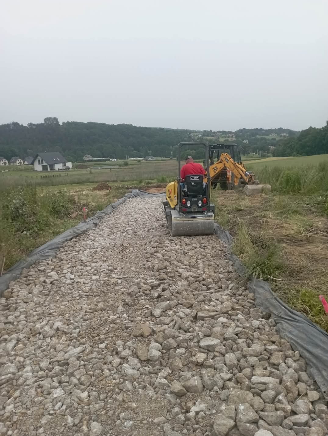 Operator w czerwonej koszuli obsługuje walec wibracyjny na drodze z tłucznia, w tle koparka JCB i krajobraz wiejski.