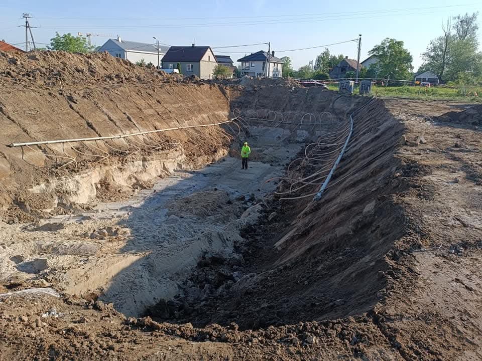 Głęboki wykop w ziemi z rurami drenażowymi na zboczu, pracownik w odblaskowej kamizelce na dnie wykopu, w tle zabudowa jednorodzinna.