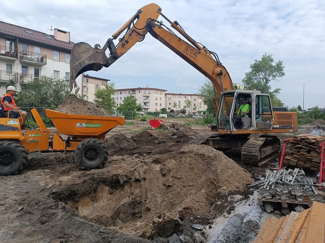Żółta koparka Liebherr 914 Litronic nabiera ziemię i wysypuje ją do wywrotki na budowie, w tle widoczne bloki mieszkalne.