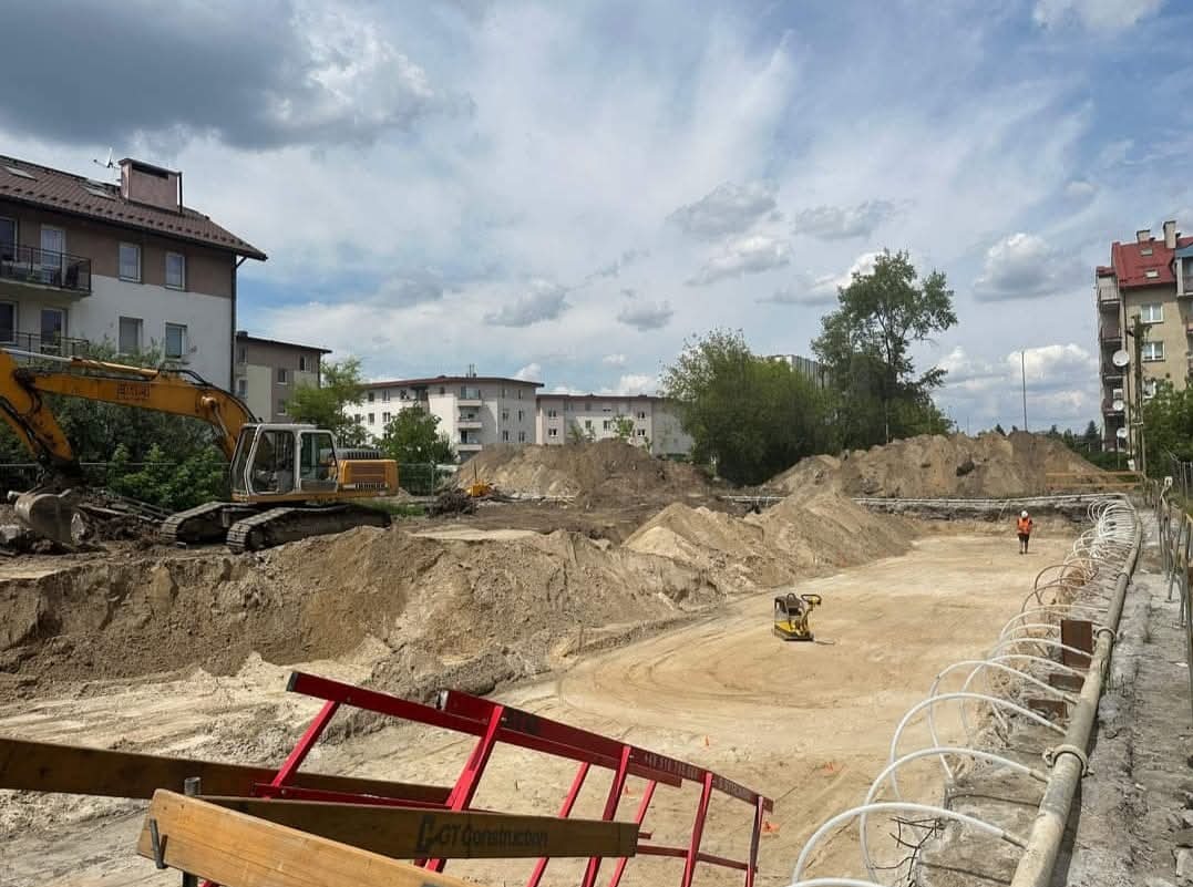 Żółta koparka na placu budowy w otoczeniu budynków mieszkalnych, widoczny ubijak wibracyjny i pracownik w pomarańczowej kamizelce, ziemia usypana w pryzmy.