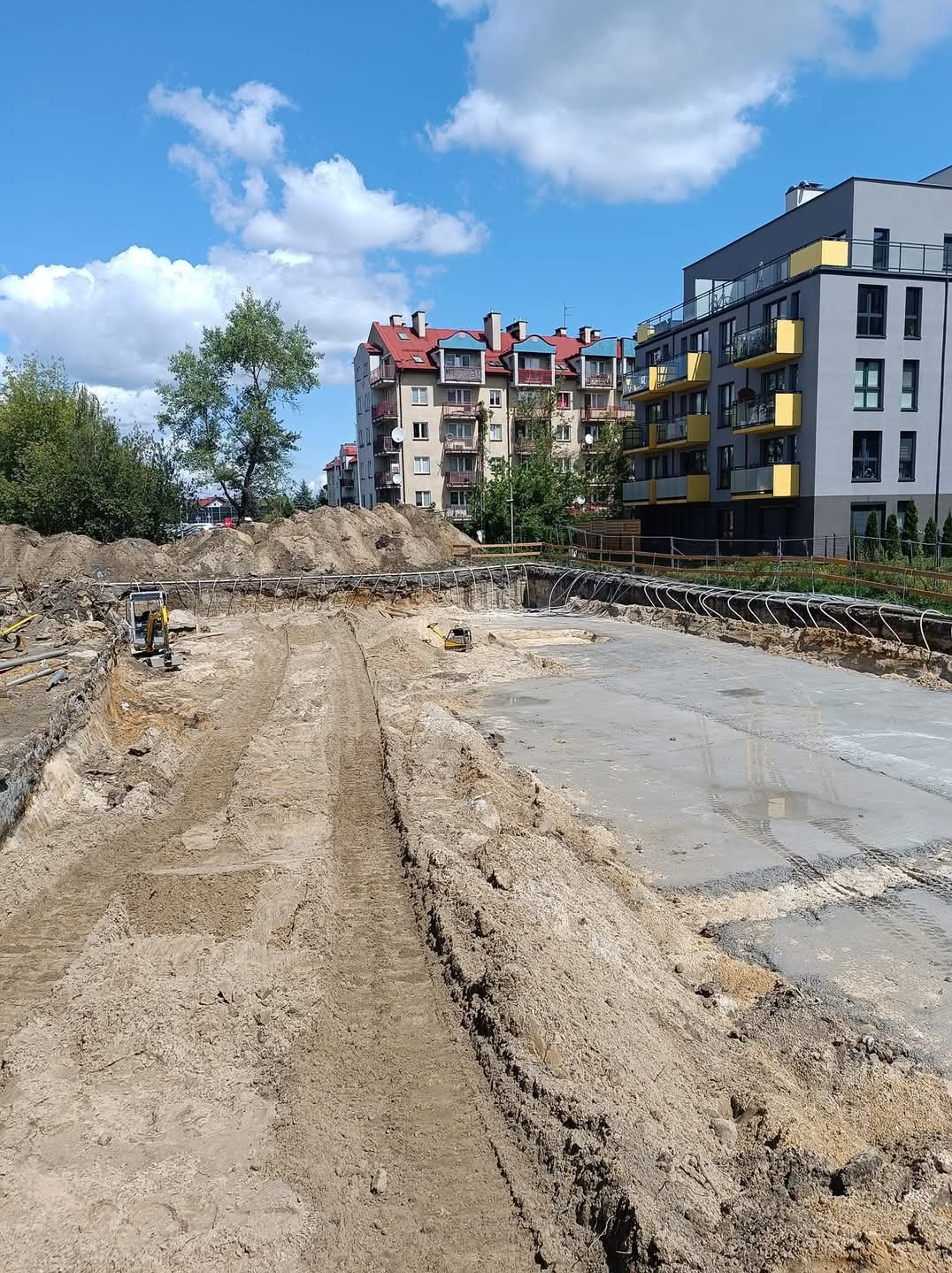 Przygotowany teren pod budowę z częściowo wylanym fundamentem, widoczne ślady opon i małe koparki, w tle budynki mieszkalne.