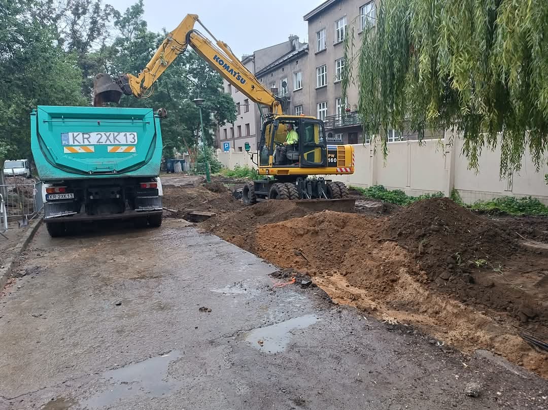 Żółta koparka Komatsu PW160 nabiera ziemię do turkusowej ciężarówki z rejestracją KR 2XK13 na tle miejskiej zabudowy i zieleni drzew