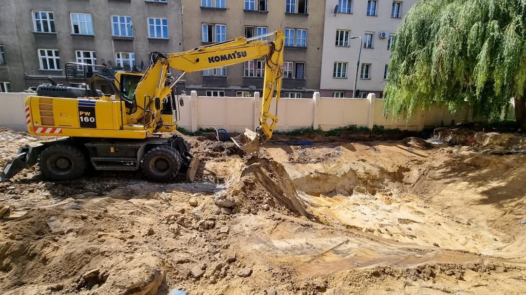 Żółta koparka Komatsu PW160 podczas prac ziemnych na placu budowy w otoczeniu miejskiej zabudowy i drzewa.