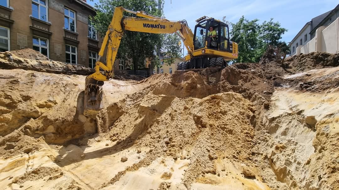 Żółta koparka Komatsu w trakcie prac ziemnych, głęboki wykop w piaszczystej glebie, w tle budynek z cegły i zieleń drzew.