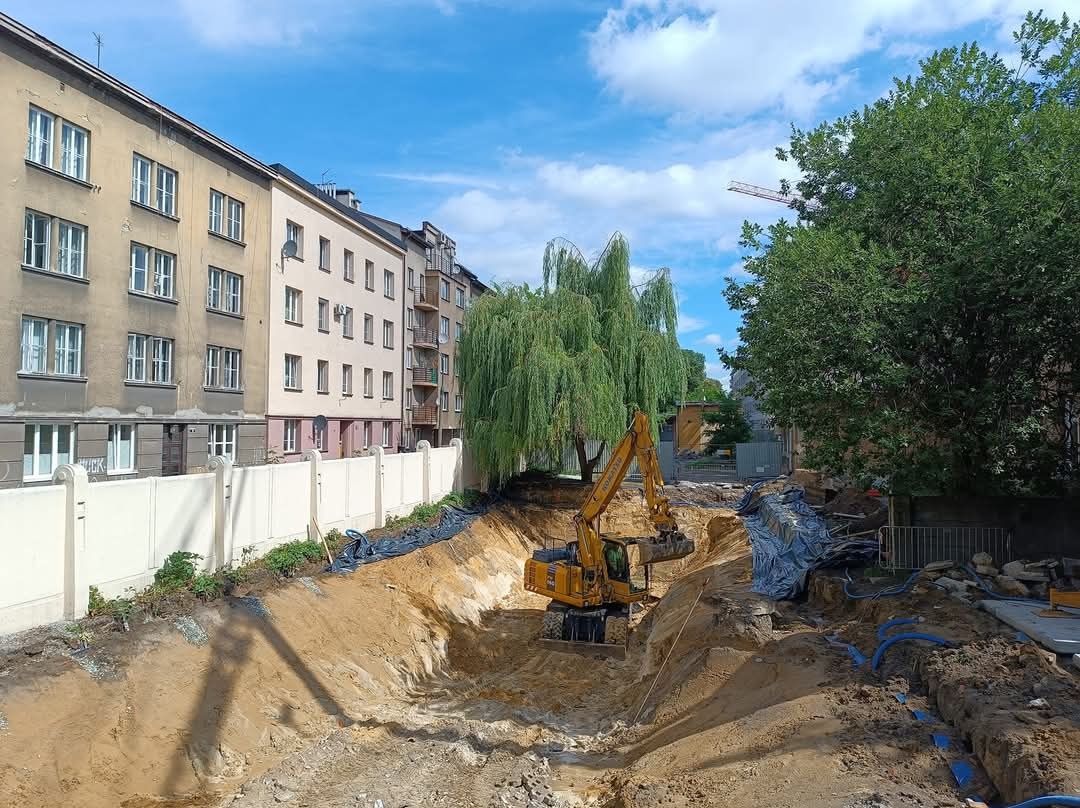 Żółta koparka w wykopie pod fundamenty, widoczne zabezpieczenia folią, w tle budynki mieszkalne i wierzba płacząca.