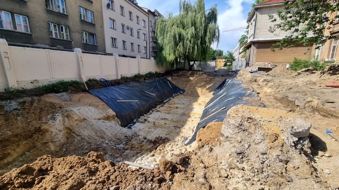 Głęboki wykop w ziemi zabezpieczony czarną folią, widoczne budynki mieszkalne w tle, częściowo pochmurne niebo.