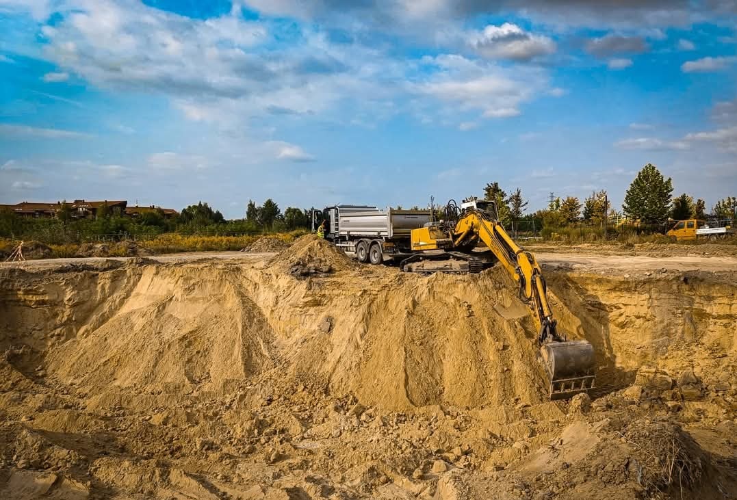 Żółta koparka na skraju wykopu, obok zaparkowana ciężarówka, w tle budynki i błękitne niebo z chmurami.