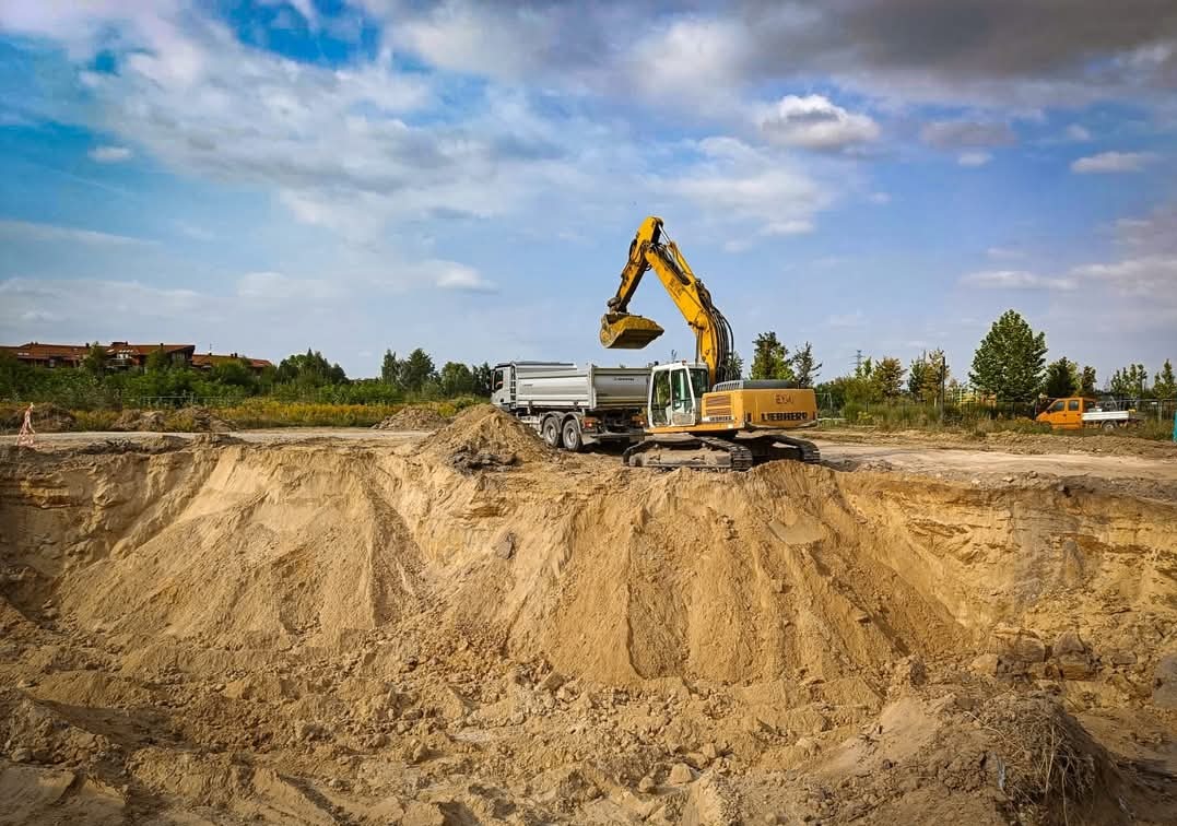 Żółto-biała koparka Liebherr ładuje urobek do wywrotki na tle wykopu w piaszczystym terenie, widoczny budynek w oddali.