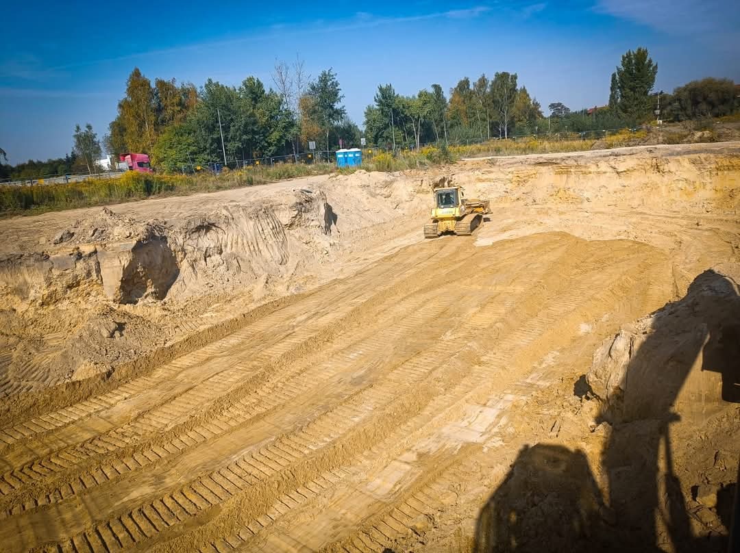 Żółta spycharka wyrównuje teren w wykopie, widoczne ślady gąsienic na piasku, w tle drzewa i przenośna toaleta.