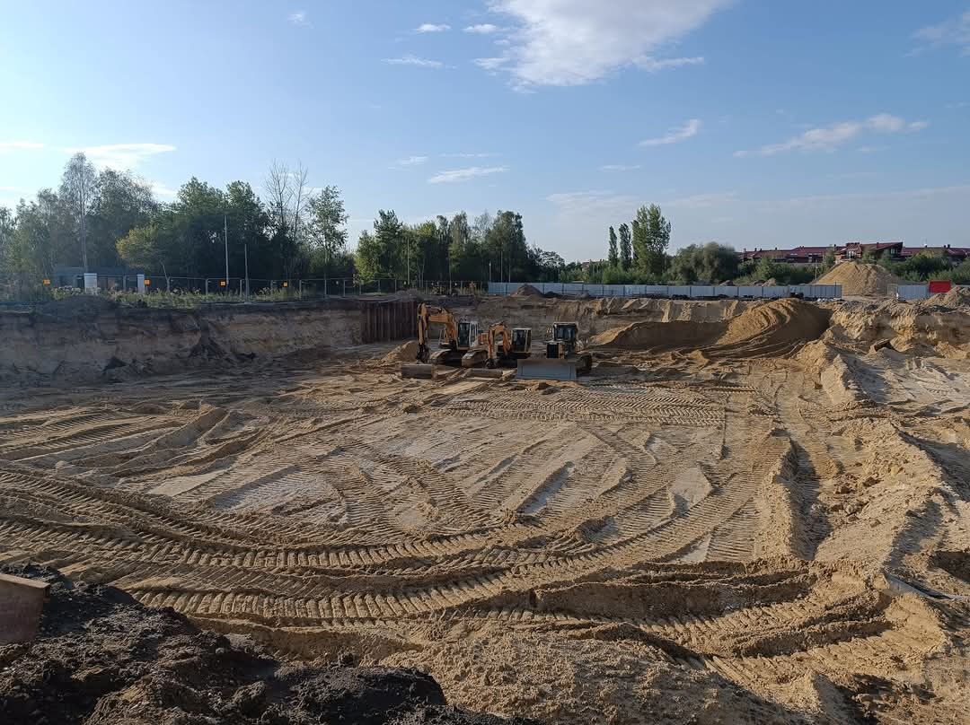 Rozległy wykop pod fundamenty z trzema maszynami budowlanymi na dnie, widoczne ślady opon na piasku, zabezpieczone ściany wykopu i drzewa w tle.