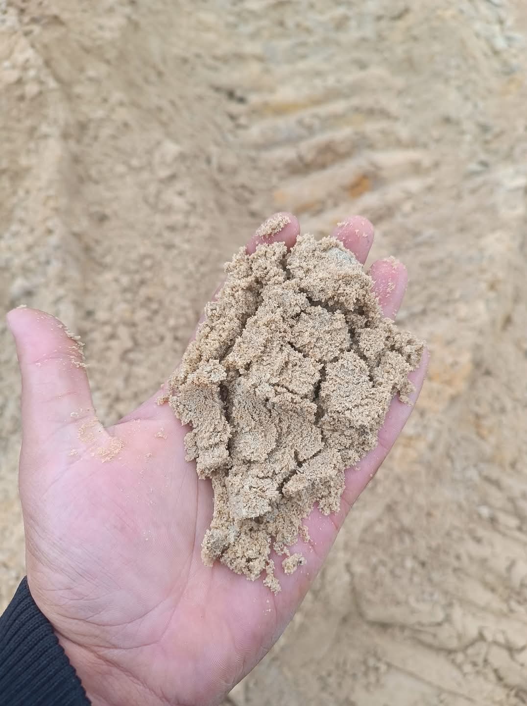 Dłoń trzymająca wilgotny, gruboziarnisty piasek w tle z widocznymi śladami opon.