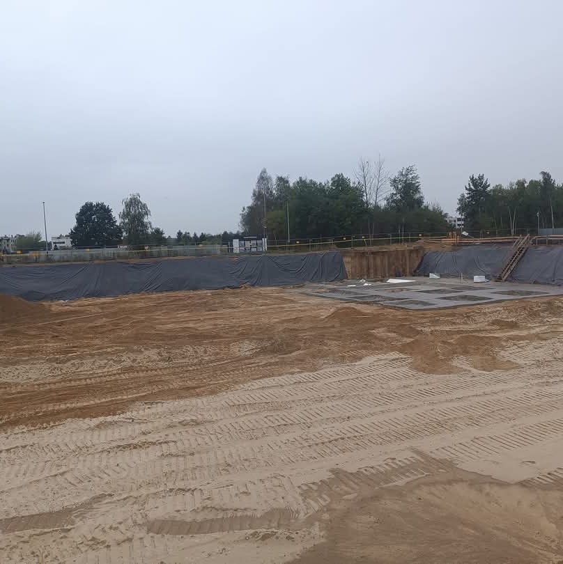 Przygotowany, rozległy wykop pod fundamenty, zabezpieczony czarną folią, z widocznymi śladami opon maszyn budowlanych na piaszczystym terenie w pochmurny dzień.