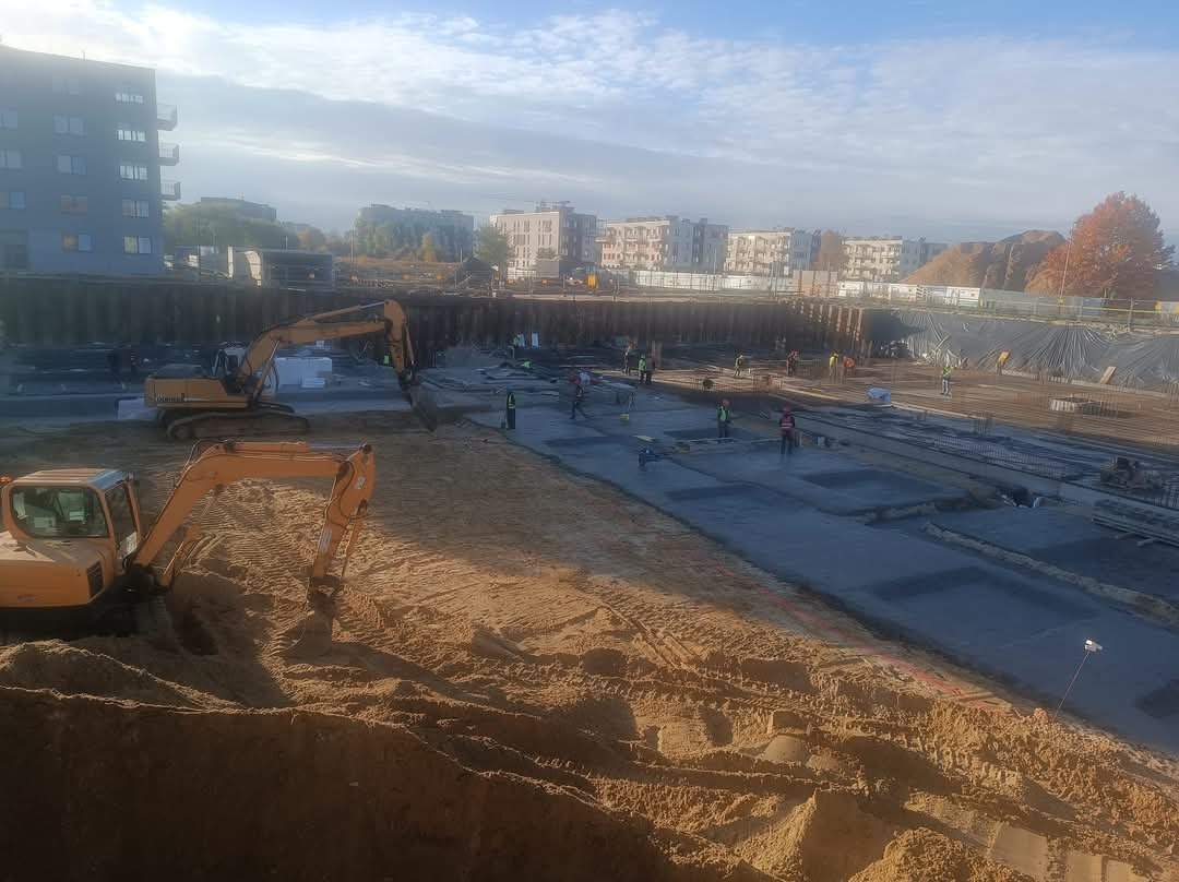 Dwa żółte koparki na placu budowy z widocznymi fundamentami i pracownikami w oddali, w tle budynki mieszkalne pod błękitnym niebem.