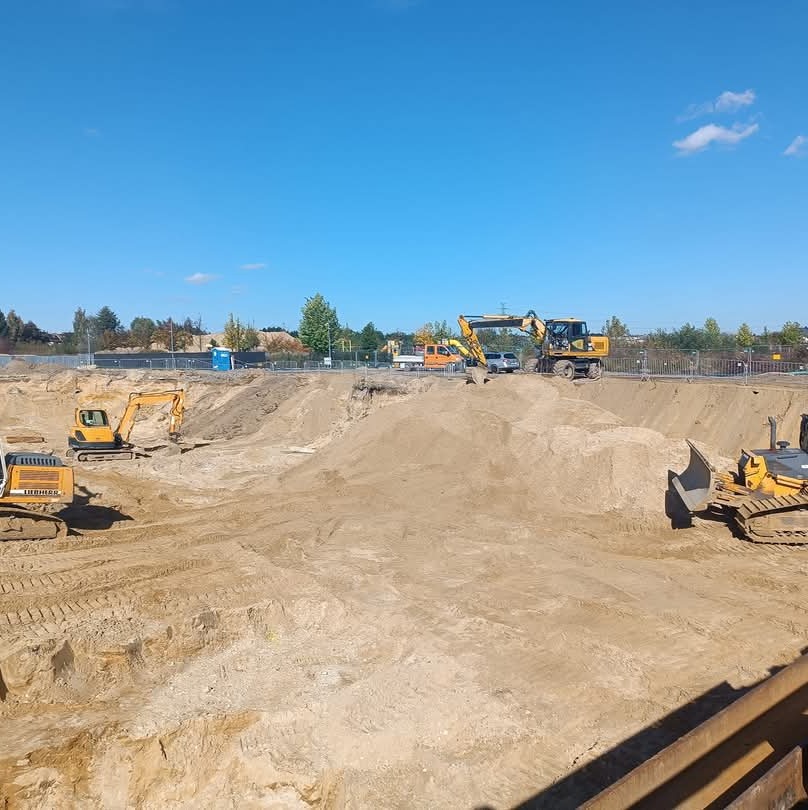 Rozległy plac budowy w słoneczny dzień z kilkoma maszynami budowlanymi, w tym koparkami i spycharką, pracującymi na nierównym terenie z piasku i ziemi. W tle widoczne drzewa i zabudowania.