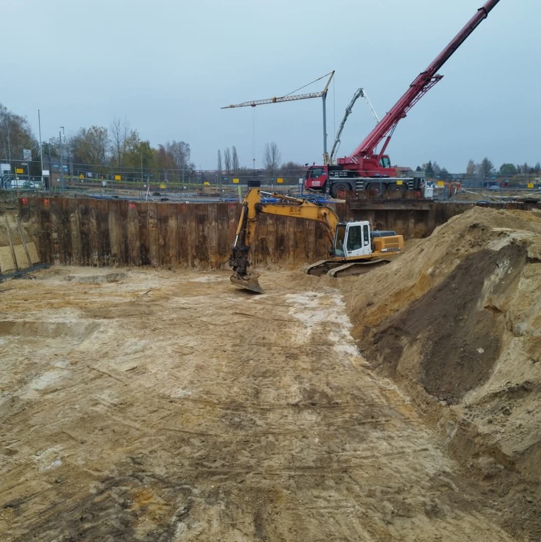 Żółta koparka na skraju wykopu zabezpieczonego stalowymi ściankami, z widocznymi żurawiami w tle, podczas prac ziemnych na placu budowy.