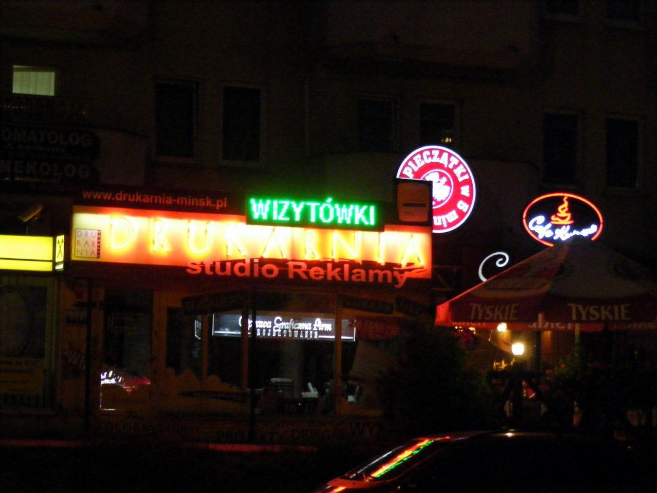 Fasada drukarni i studia reklamy w Mińsku Mazowieckim, oświetlona neonami z napisami 'Drukarnia', 'Studio Reklamy' i 'Wizytówki', widoczne również inne neony reklamowe i parasole Tyskie.