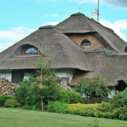 Tradycyjny dom kryty strzechą z okrągłymi oknami, z zapasem drewna ułożonym przy ścianie, otoczony zielenią trawnika i drzew.