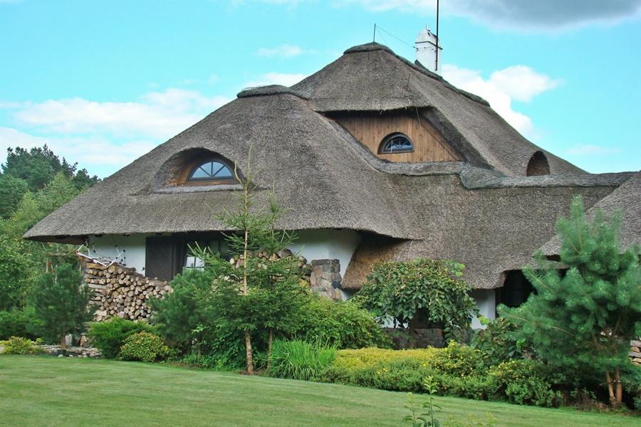 Tradycyjny dom kryty strzechą z okrągłymi oknami, z zapasem drewna ułożonym przy ścianie, otoczony zielenią trawnika i drzew.