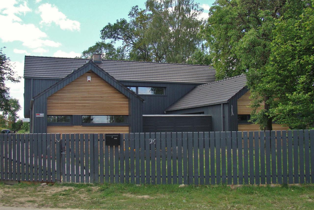 Nowoczesny dom jednorodzinny z szarą drewnianą elewacją i elementami z naturalnego drewna, widoczny zza grafitowego płotu sztachetowego z numerem 24.