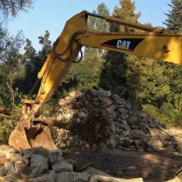 Żółta koparka CAT z łyżką nabiera kamienie i gałęzie na tle zieleni drzew, przygotowując teren pod prace ogrodowe lub budowlane.
