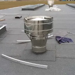 Montaż komina wentylacyjnego na dachu pokrytym papą, widoczne narzędzia, fragmenty papy, i elementy obróbki blacharskiej, w tle świetliki dachowe.