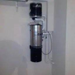 Centralny odkurzacz zainstalowany w pomieszczeniu technicznym, widoczne rury i jednostka centralna z metalowym zbiornikiem i czarnymi elementami, zamontowana na ścianie w rogu pomieszczenia.