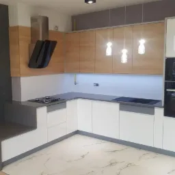 Nowoczesna kuchnia w kształcie litery L z białymi frontami szafek dolnych i drewnopodobnymi górnymi, szary blat, płyta gazowa i indukcyjna, piekarnik i mikrofalówka w zabudowie, marmurowa podłoga...