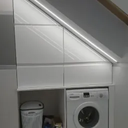 Biała zabudowa meblowa pod skosem dachu z oświetleniem LED, pralka i kosz na pranie wkomponowane w aranżację.