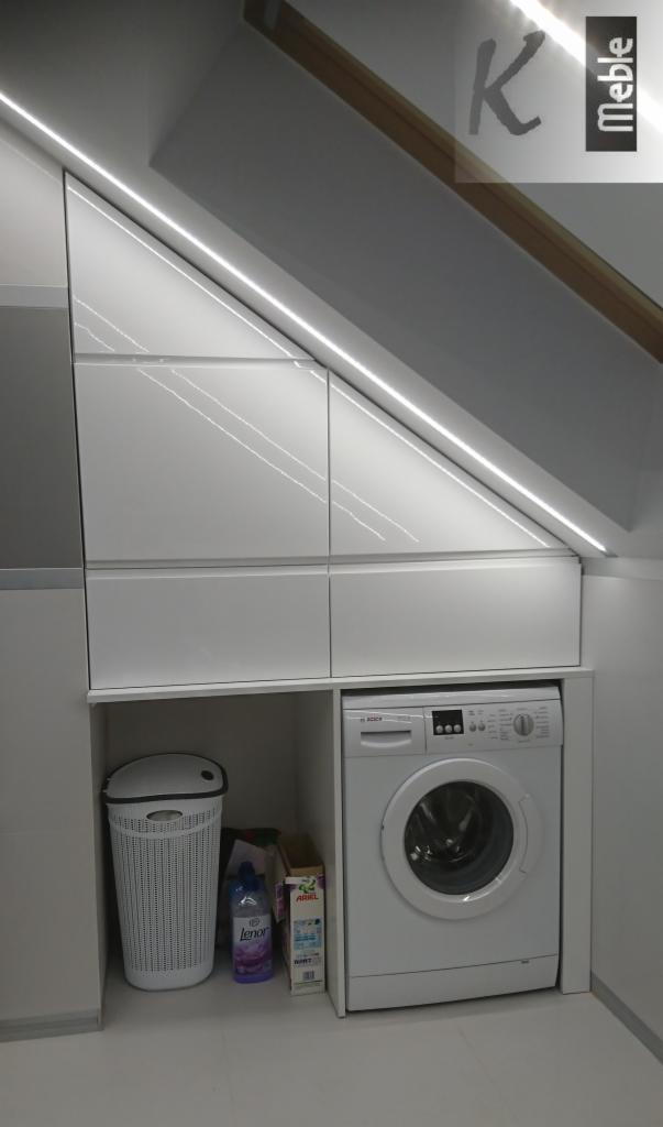 Biała zabudowa meblowa pod skosem dachu z oświetleniem LED, pralka i kosz na pranie wkomponowane w aranżację.