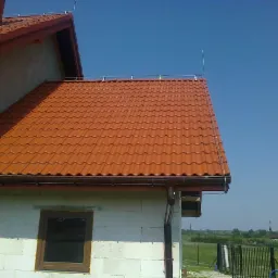 Pomarańczowa dachówka ceramiczna na domu w trakcie budowy, z widoczną instalacją odgromową, mocowaną do dachu i ściany budynku.