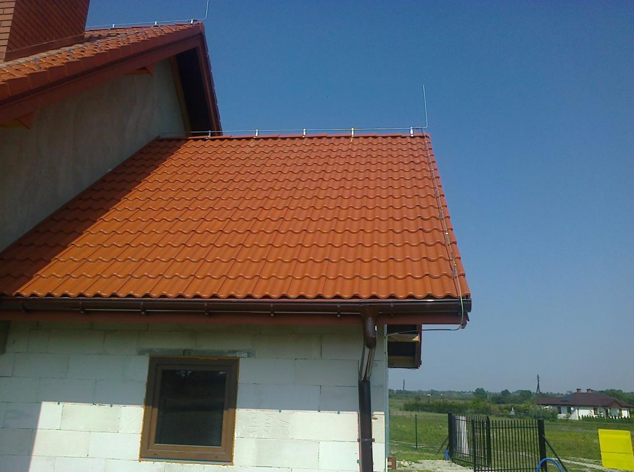 Pomarańczowa dachówka ceramiczna na domu w trakcie budowy, z widoczną instalacją odgromową, mocowaną do dachu i ściany budynku.