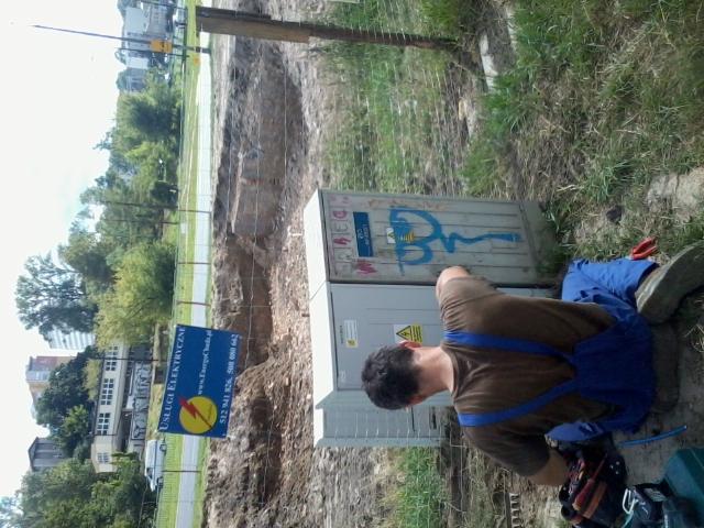 Elektryk w niebieskich spodniach roboczych i brązowej koszulce pracuje przy skrzynce elektrycznej na budowie, obok widoczny znak z ofertą usług elektrycznych.