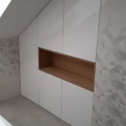 Biała szafa wnękowa pod skosem dachu z drewnianą wnęką, nowoczesna łazienka z geometrycznymi płytkami.
