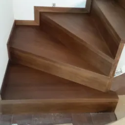 Wykończone schody drewniane z ciemnego drewna, widok z góry, widoczna biała ściana i zabezpieczone podłoże kartonem.