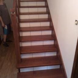 Schody drewniane Meble Adrian - Klatka schodowa z drewnianymi stopniami i białymi podstopnicami z płytek, widok z dołu, osoba w tle patrzy w górę.