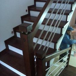 Schody drewniane Meble Adrian - Ciemnobrązowe schody z drewnianymi stopniami i białymi podstopnicami, balustrada z drewnianymi słupkami i metalowymi prętami, widoczny pracownik w niebieskiej kurtce.