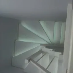 Spiralne, białe schody z podświetleniem LED wzdłuż stopni, widok z dołu, minimalistyczny design.