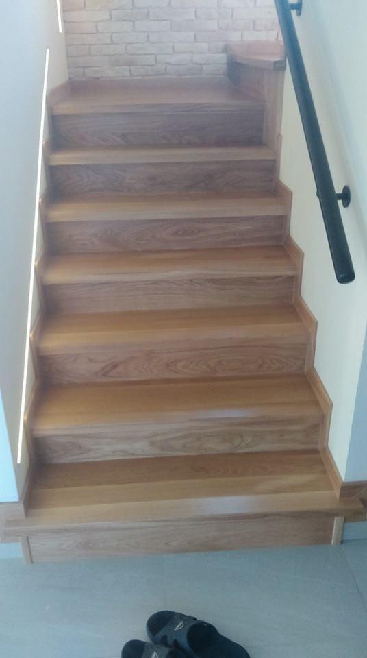 Jasne, drewniane schody z czarną poręczą, oświetleniem LED przy ścianie i ceglanym wykończeniem ściany w tle; na podłodze leżą klapki.