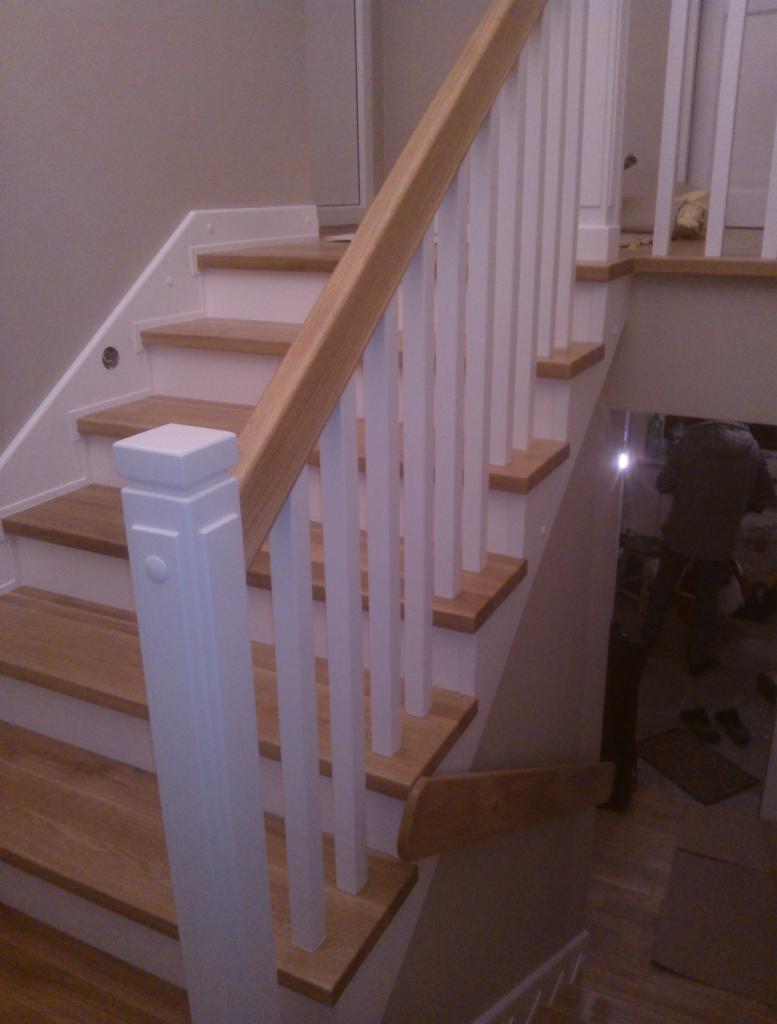 Jasne schody z drewnianymi stopniami i białymi balustradami, widoczne odbicie osoby w szybie na półpiętrze.