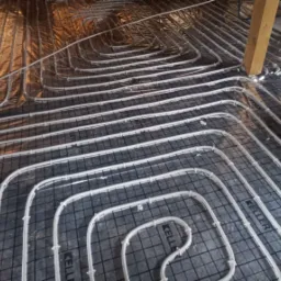 Rozłożone rury ogrzewania podłogowego na folii izolacyjnej z rastrem, widoczny fragment drewnianej konstrukcji stropu.