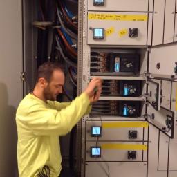 Monterzy.eu - Elektryk w żółtej koszulce, klęcząc przed otwartą szafą elektryczną, używa szczypiec do pracy przy przewodach. Widoczne liczne kable, mierniki i wyłączniki w szafie. Na pasie narzędziowym montera...