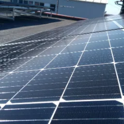 Rząd paneli fotowoltaicznych na płaskim dachu z widocznymi mocowaniami i odblaskami słońca.