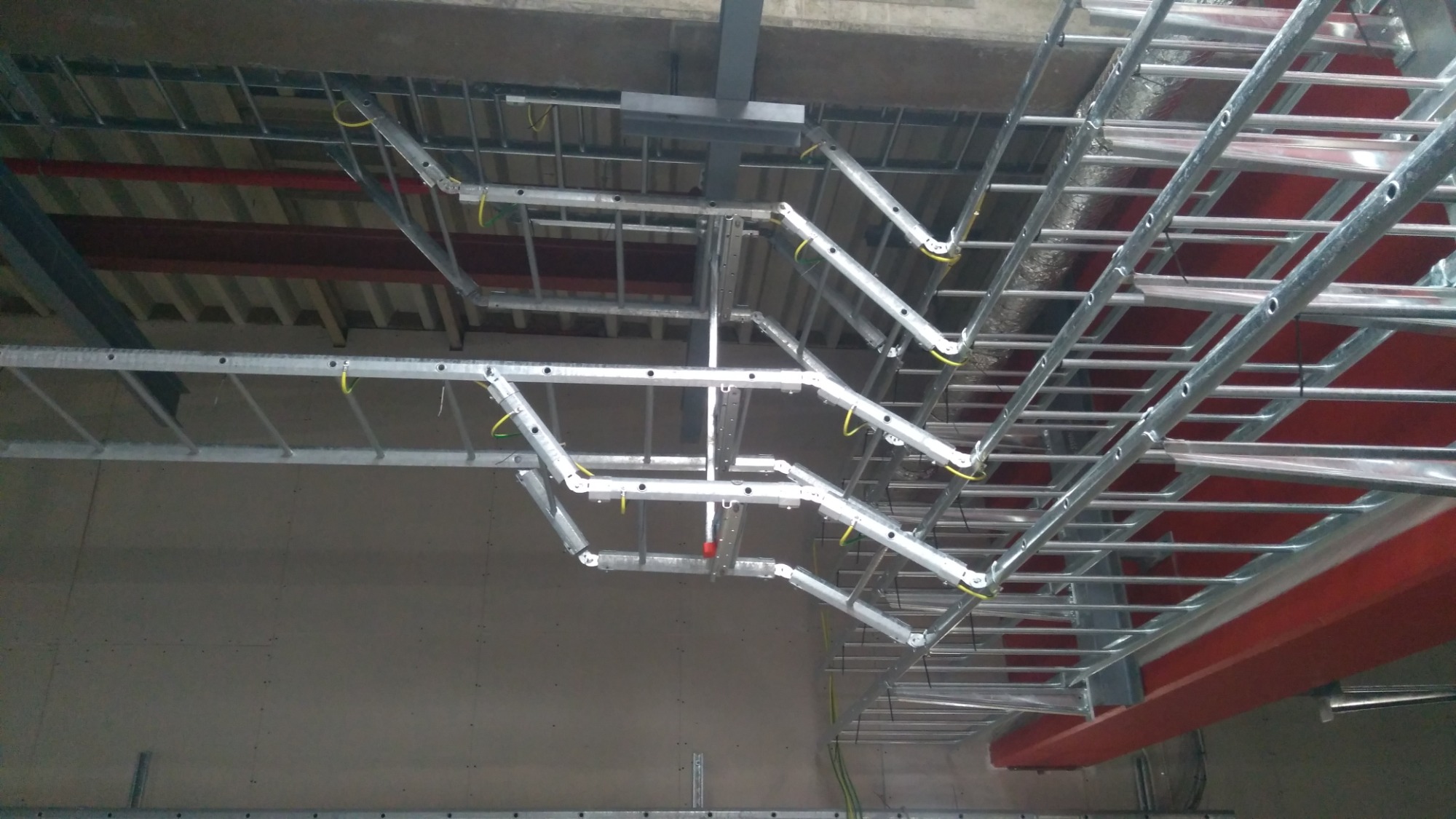 Metalowe korytka kablowe zamontowane pod sufitem, z widocznymi przewodami w kolorze żółto-zielonym, układane wzdłuż belek konstrukcyjnych.