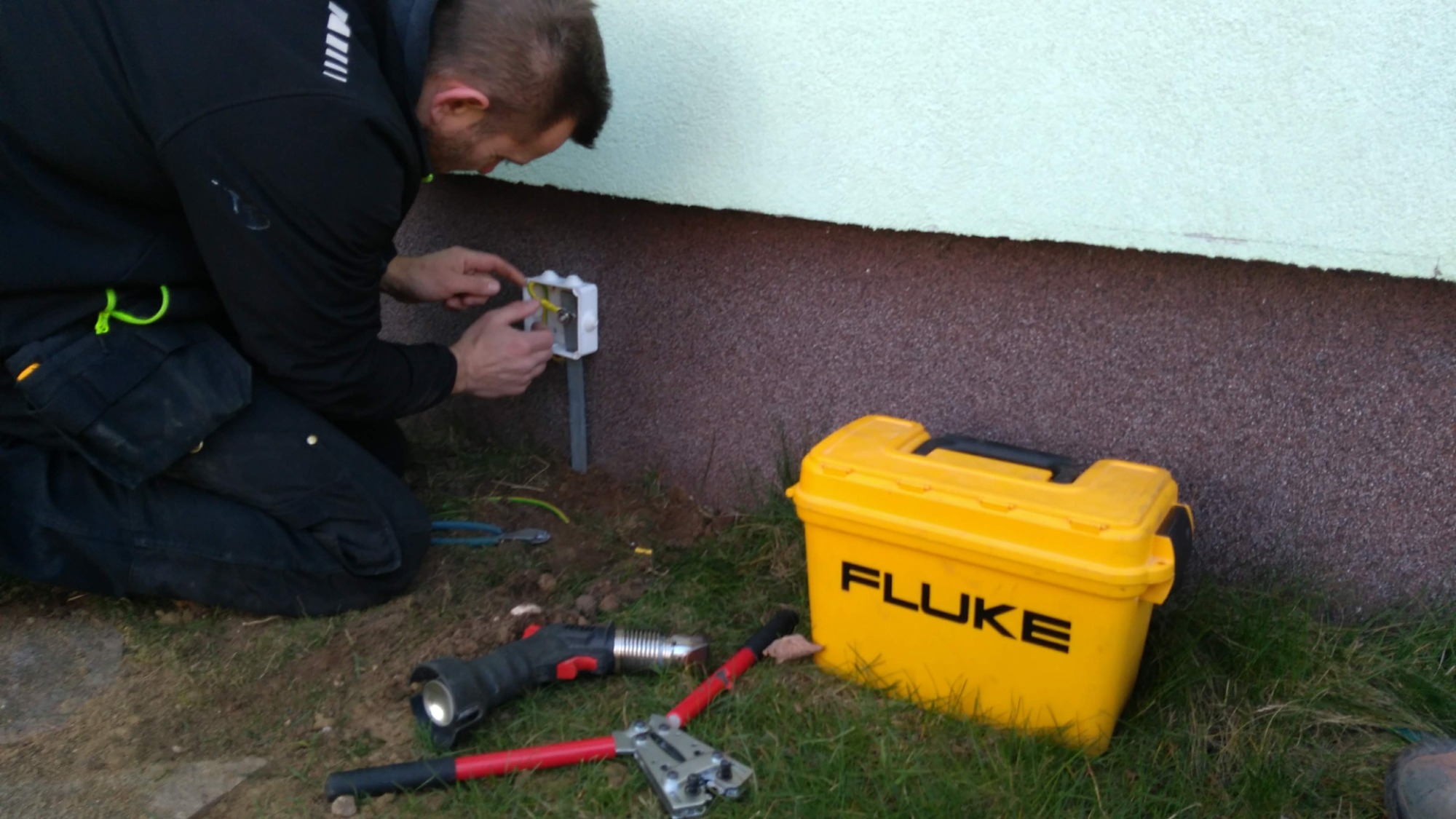 Elektryk podłącza przewody w puszce elektrycznej na elewacji budynku, obok leżą narzędzia i żółta skrzynka narzędziowa firmy Fluke.