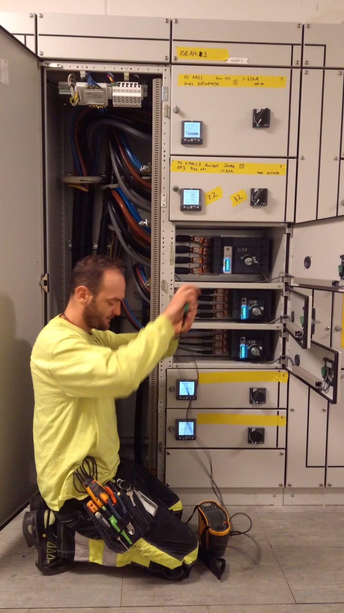 Elektryk w żółtej koszulce, klęcząc przed otwartą szafą elektryczną, używa szczypiec do pracy przy przewodach. Widoczne liczne kable, mierniki i wyłączniki w szafie. Na pasie narzędziowym montera...