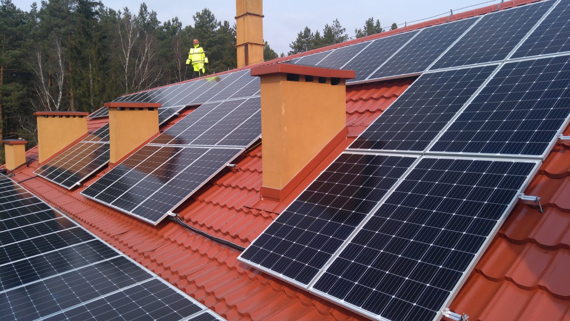 Instalacja paneli słonecznych na dachu z czerwonej dachówki, widoczny pracownik w odblaskowym kombinezonie stojący na dachu w tle lasu.