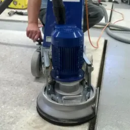 Szlifowanie posadzki przemysłowej za pomocą profesjonalnej szlifierki Anser-330 z odkurzaczem przemysłowym, widoczny operator w trakcie pracy.