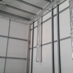 Izolowane kanały wentylacyjne w hali przemysłowej, widoczne metalowe ramy konstrukcyjne i białe ściany z paneli.