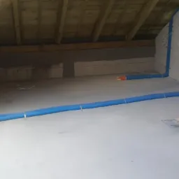 Montaż instalacji wentylacyjnej z niebieskich rur karbowanych w surowym wnętrzu poddasza, widoczne elementy konstrukcji drewnianej i ściany z betonu komórkowego.